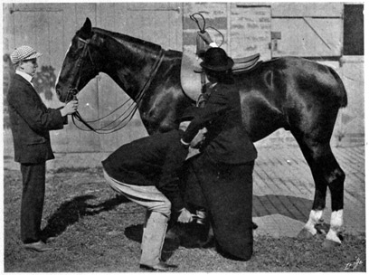 Three people standing near a horse. A groom holding the horse's head, a man crouching to lift the woman who has one hand on his shoulder and the other on the saddle